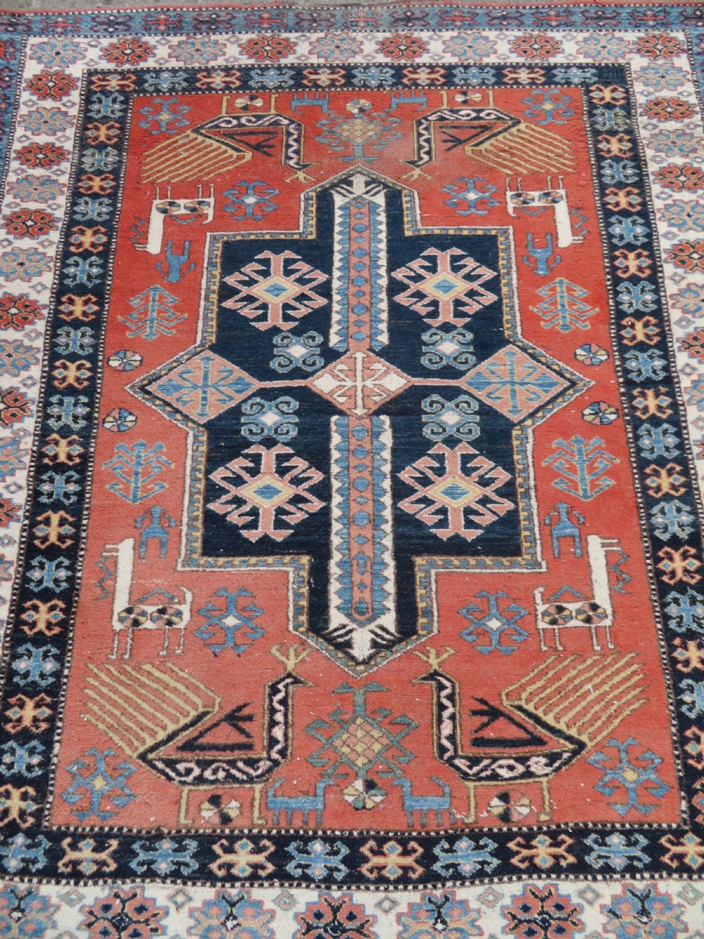 A handwoven rectangular wool rug featuring a central navy blue stepped medallion and a terracotta field decorated with peacock and animal motifs. View III