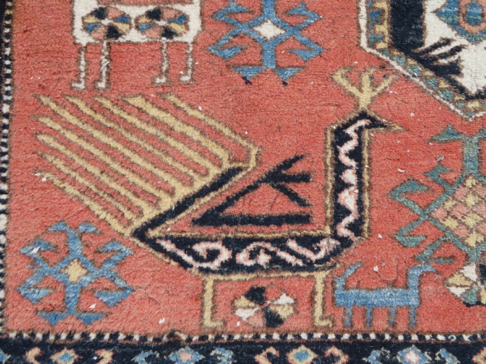 A handwoven rectangular wool rug featuring a central navy blue stepped medallion and a terracotta field decorated with peacock and animal motifs. View IV