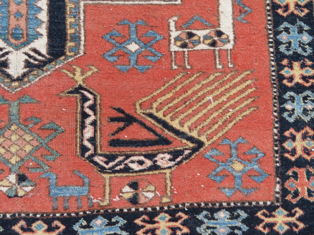 A handwoven rectangular wool rug featuring a central navy blue stepped medallion and a terracotta field decorated with peacock and animal motifs. View V