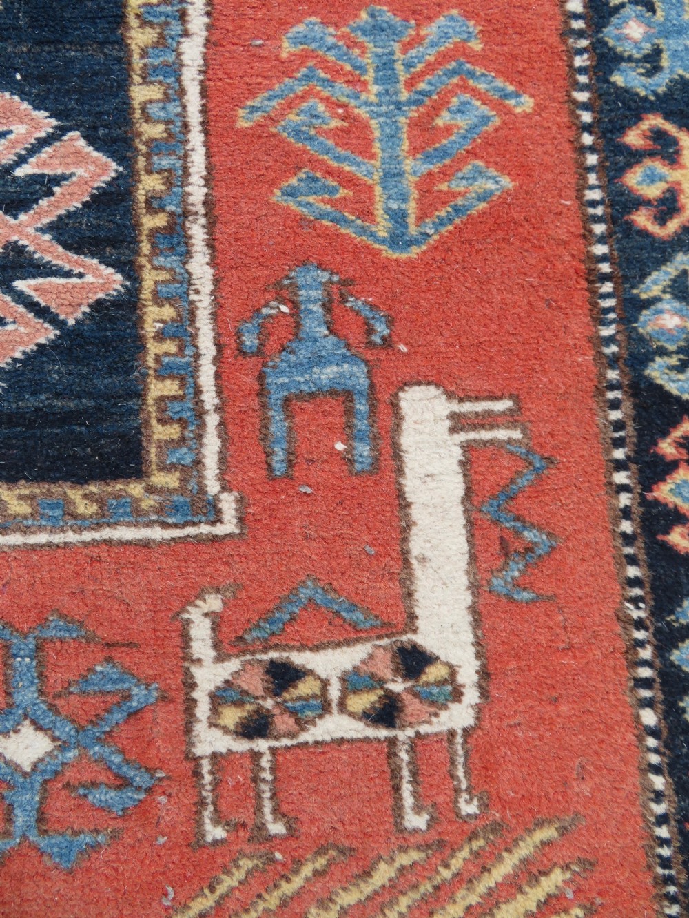 A handwoven rectangular wool rug featuring a central navy blue stepped medallion and a terracotta field decorated with peacock and animal motifs. View VI