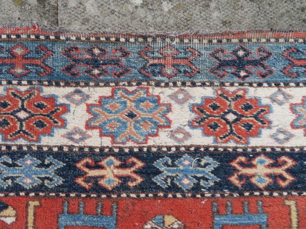 A handwoven rectangular wool rug featuring a central navy blue stepped medallion and a terracotta field decorated with peacock and animal motifs. View IX