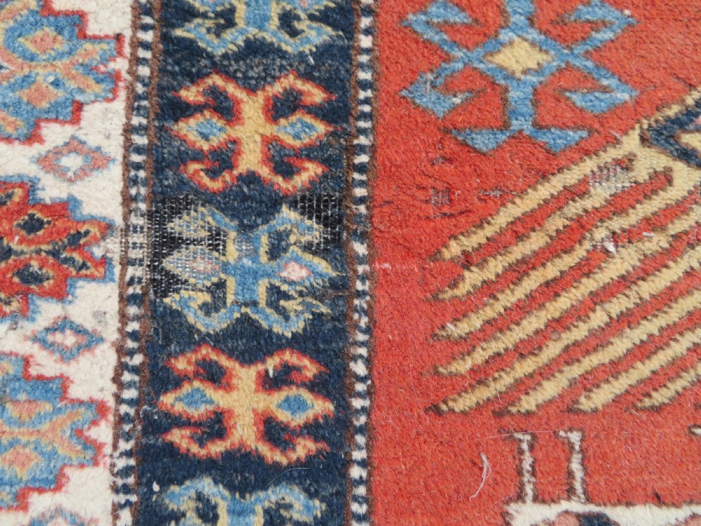 A handwoven rectangular wool rug featuring a central navy blue stepped medallion and a terracotta field decorated with peacock and animal motifs. View XII