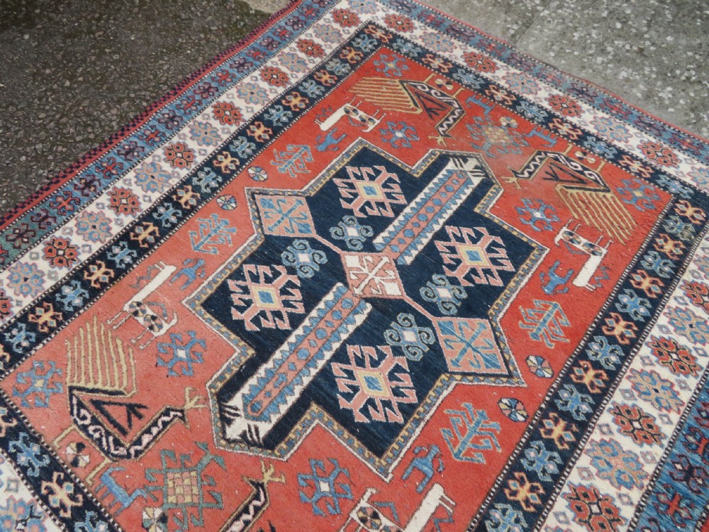 A handwoven rectangular wool rug featuring a central navy blue stepped medallion and a terracotta field decorated with peacock and animal motifs. View XIV