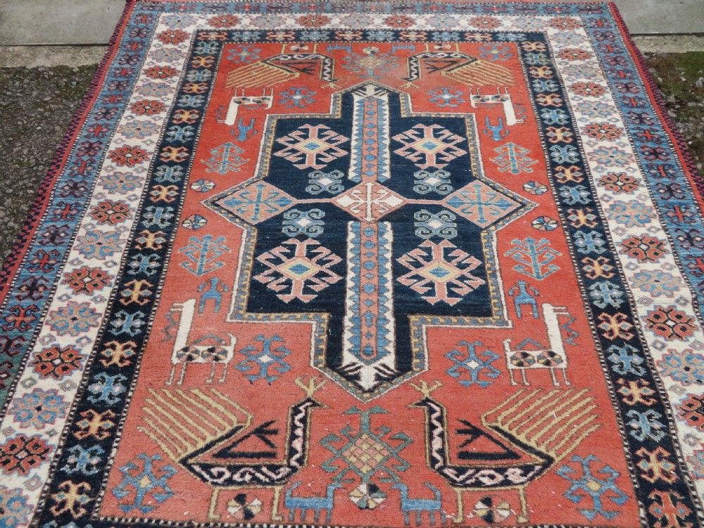 A handwoven rectangular wool rug featuring a central navy blue stepped medallion and a terracotta field decorated with peacock and animal motifs. View XV