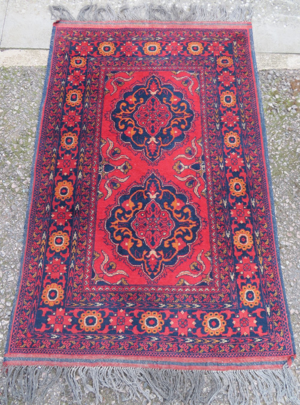 A rectangular hand-knotted wool rug featuring two central dark blue geometric medallions on a deep red field framed by decorated floral borders and fringe ends.