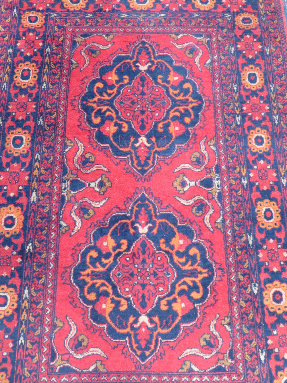 A rectangular hand-knotted wool rug featuring two central dark blue geometric medallions on a deep red field framed by decorated floral borders and fringe ends. View II