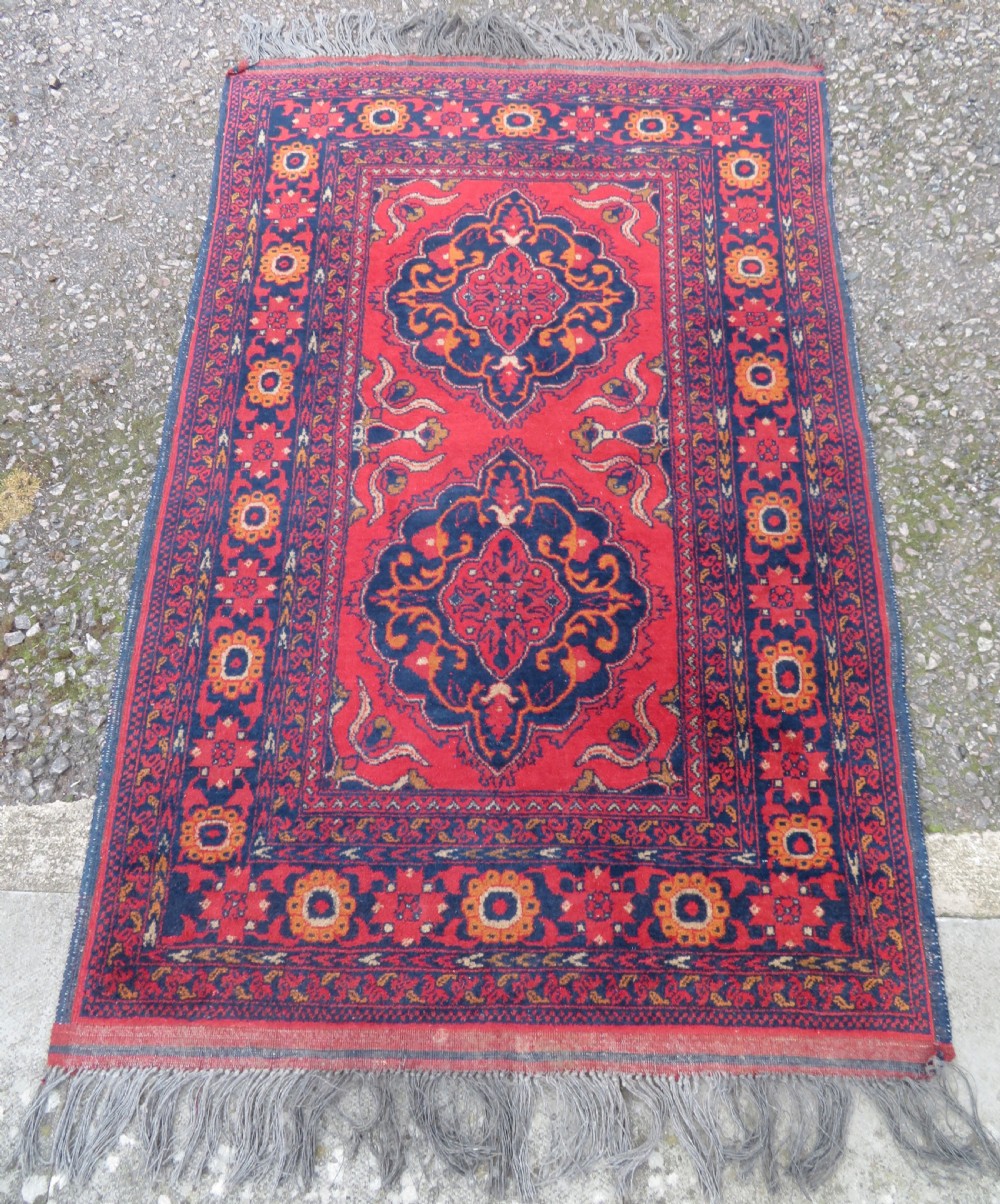A rectangular hand-knotted wool rug featuring two central dark blue geometric medallions on a deep red field framed by decorated floral borders and fringe ends. View III