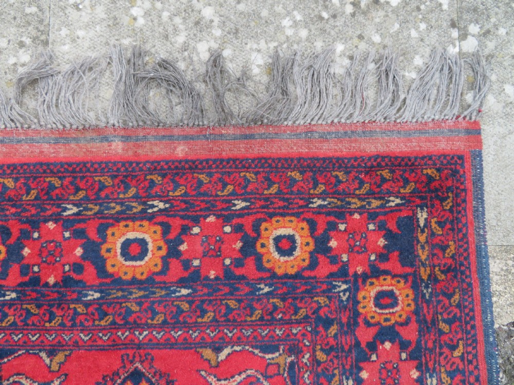 A rectangular hand-knotted wool rug featuring two central dark blue geometric medallions on a deep red field framed by decorated floral borders and fringe ends. View V