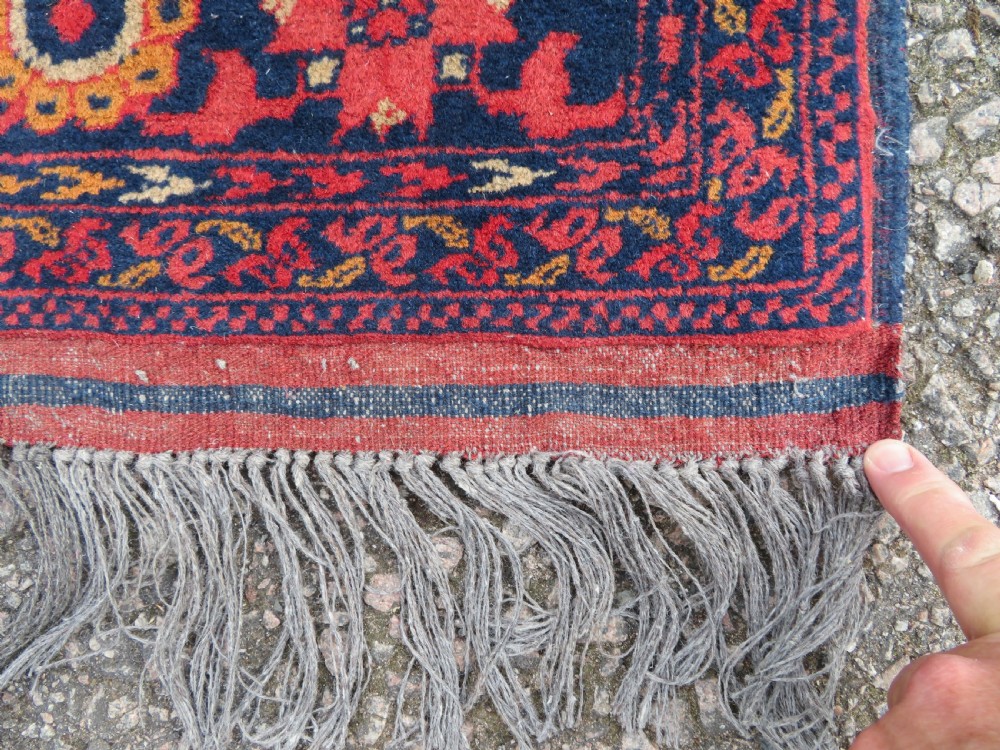 A rectangular hand-knotted wool rug featuring two central dark blue geometric medallions on a deep red field framed by decorated floral borders and fringe ends. View X
