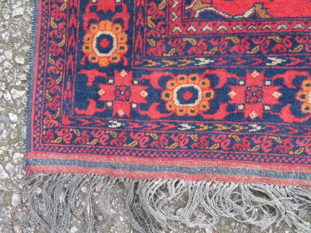 A rectangular hand-knotted wool rug featuring two central dark blue geometric medallions on a deep red field framed by decorated floral borders and fringe ends. View XI