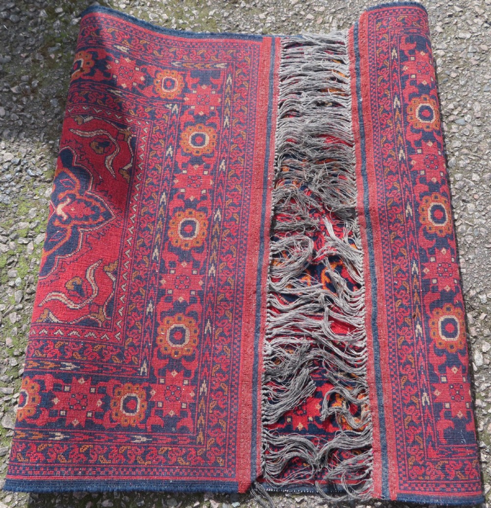 A rectangular hand-knotted wool rug featuring two central dark blue geometric medallions on a deep red field framed by decorated floral borders and fringe ends. View XII