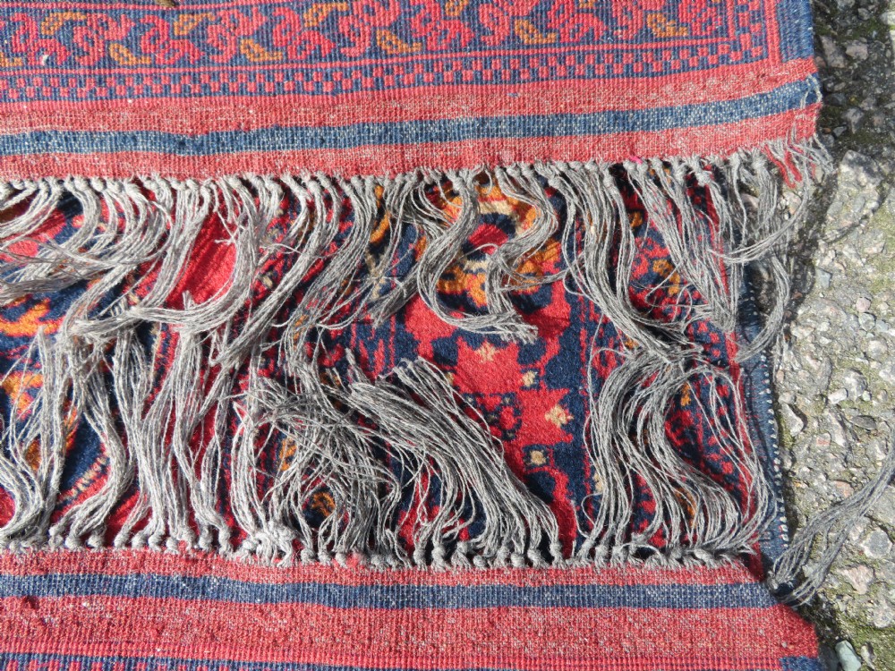 A rectangular hand-knotted wool rug featuring two central dark blue geometric medallions on a deep red field framed by decorated floral borders and fringe ends. View XIII