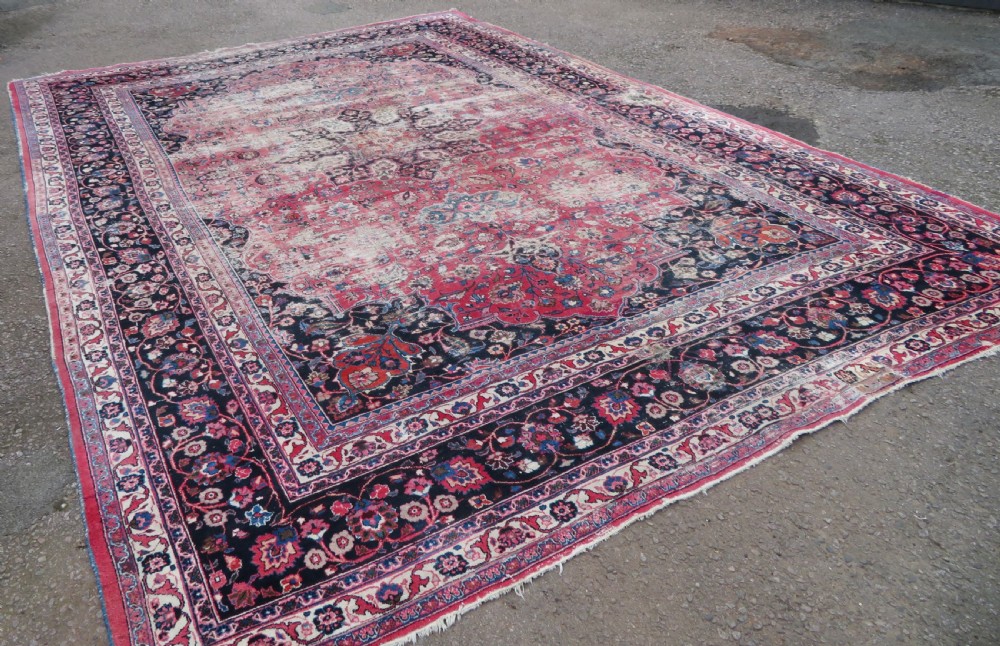 A large hand-knotted Persian Sarouk wool carpet featuring a distressed rose red field, central floral medallion, and deep navy blue floral borders.