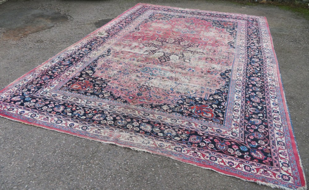 A large hand-knotted Persian Sarouk wool carpet featuring a distressed rose red field, central floral medallion, and deep navy blue floral borders. View IV