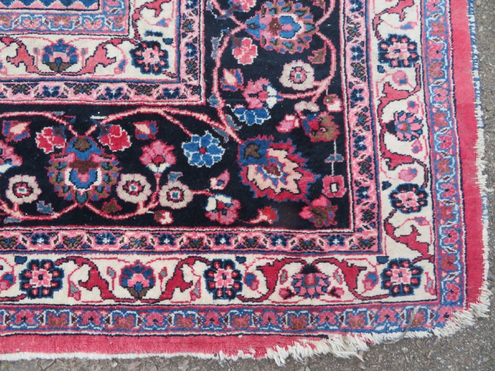 A large hand-knotted Persian Sarouk wool carpet featuring a distressed rose red field, central floral medallion, and deep navy blue floral borders. View VI