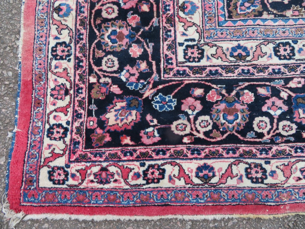 A large hand-knotted Persian Sarouk wool carpet featuring a distressed rose red field, central floral medallion, and deep navy blue floral borders. View VII