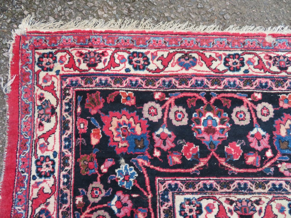 A large hand-knotted Persian Sarouk wool carpet featuring a distressed rose red field, central floral medallion, and deep navy blue floral borders. View VIII