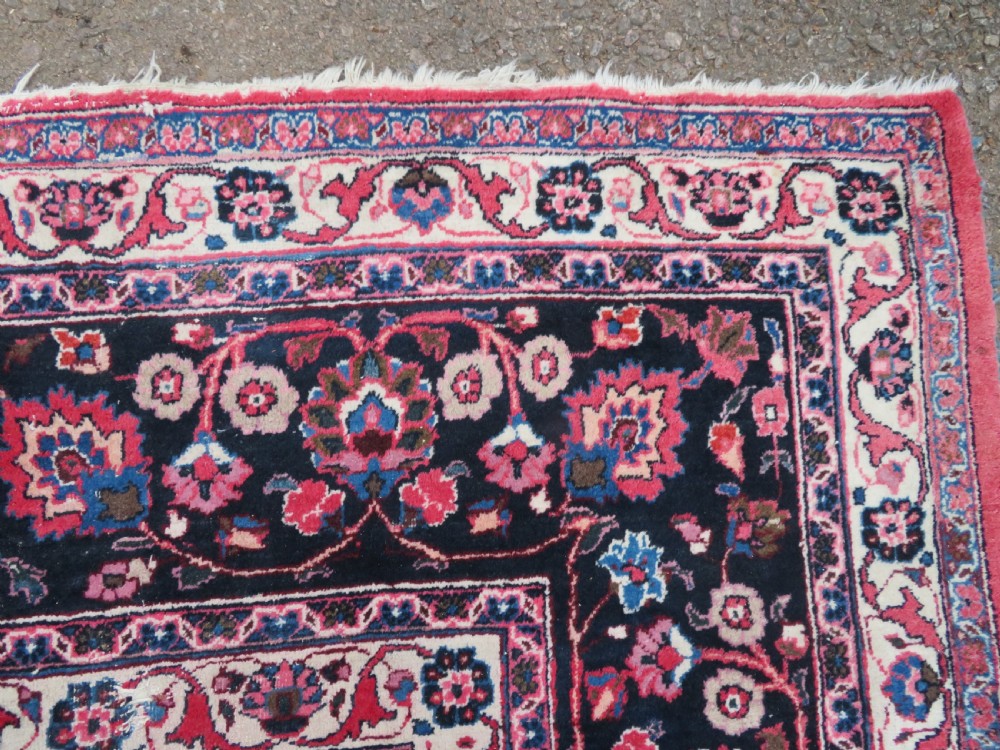 A large hand-knotted Persian Sarouk wool carpet featuring a distressed rose red field, central floral medallion, and deep navy blue floral borders. View IX