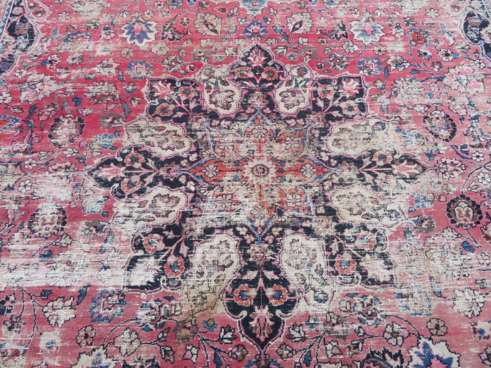 A large hand-knotted Persian Sarouk wool carpet featuring a distressed rose red field, central floral medallion, and deep navy blue floral borders. View XII