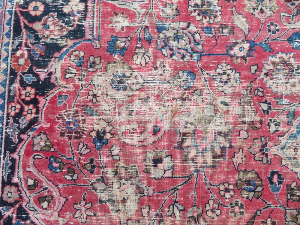 A large hand-knotted Persian Sarouk wool carpet featuring a distressed rose red field, central floral medallion, and deep navy blue floral borders. View XV