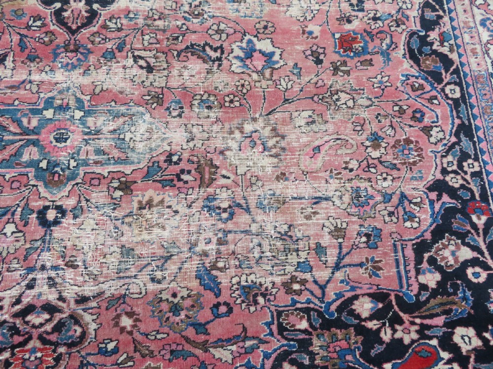 A large hand-knotted Persian Sarouk wool carpet featuring a distressed rose red field, central floral medallion, and deep navy blue floral borders. View XVI