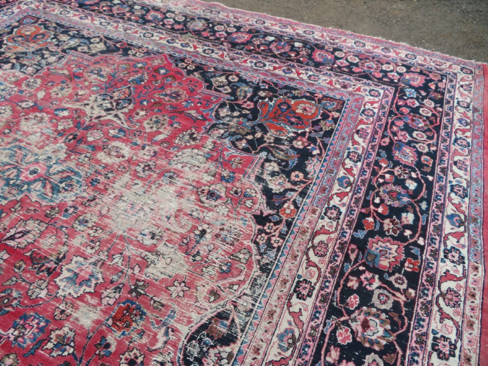 A large hand-knotted Persian Sarouk wool carpet featuring a distressed rose red field, central floral medallion, and deep navy blue floral borders. View XIX