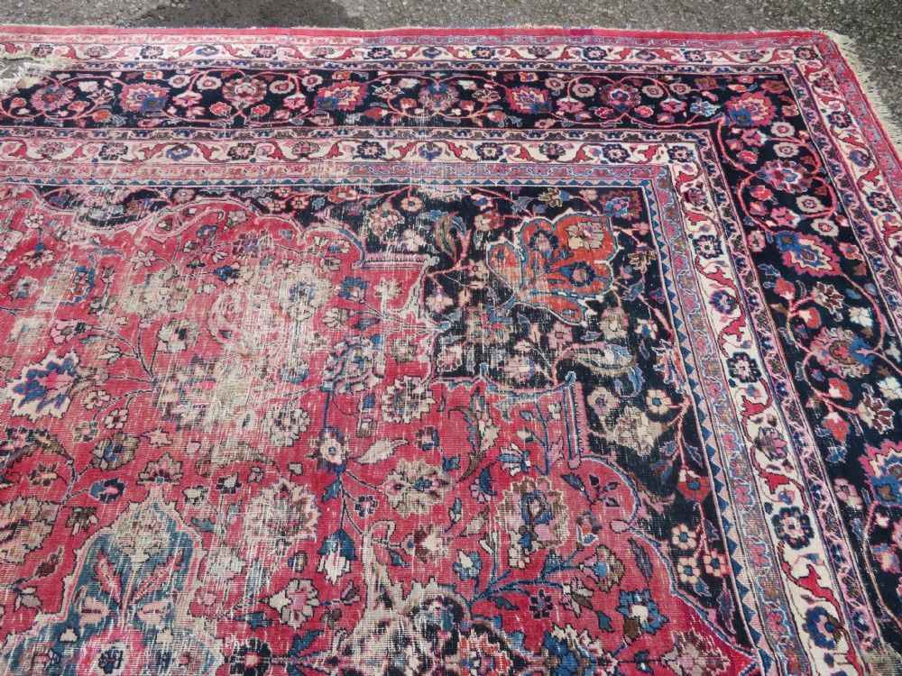 A large hand-knotted Persian Sarouk wool carpet featuring a distressed rose red field, central floral medallion, and deep navy blue floral borders. View XXI