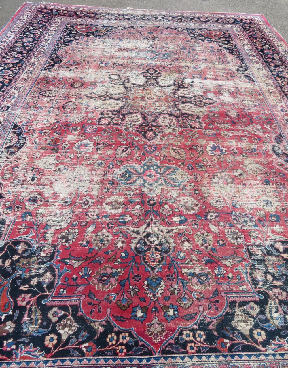A large hand-knotted Persian Sarouk wool carpet featuring a distressed rose red field, central floral medallion, and deep navy blue floral borders. View XXV