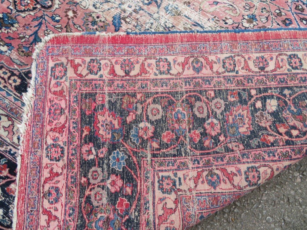 A large hand-knotted Persian Sarouk wool carpet featuring a distressed rose red field, central floral medallion, and deep navy blue floral borders. View XXX