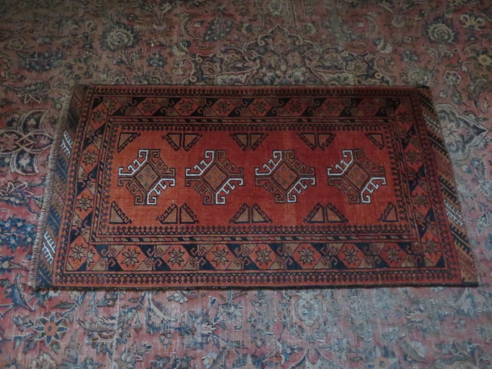 A hand-knotted Afghan wool rug featuring a rust-red field decorated with four diamond latch-hook medallions and geometric borders. View IV