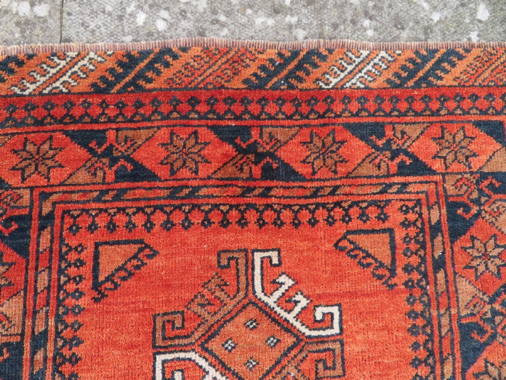 A hand-knotted Afghan wool rug featuring a rust-red field decorated with four diamond latch-hook medallions and geometric borders. View VI