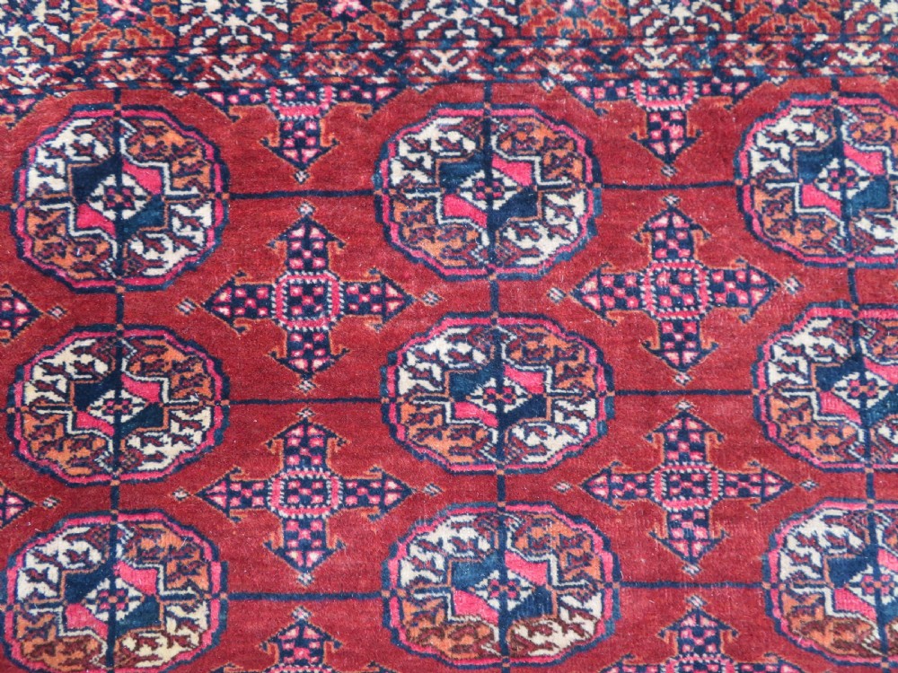 A rectangular hand-knotted Tekke Turkmen wool rug featuring three columns of octagonal gol medallions on a deep red field with geometric borders and fringed ends. View V