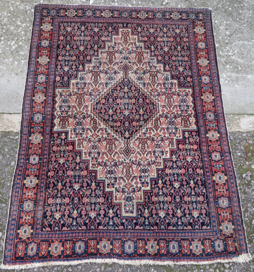 An antique Kurdish Senneh hand-knotted wool rug featuring a central stepped diamond medallion surrounded by detailed repeating motifs in navy blue, terracotta red, and cream.