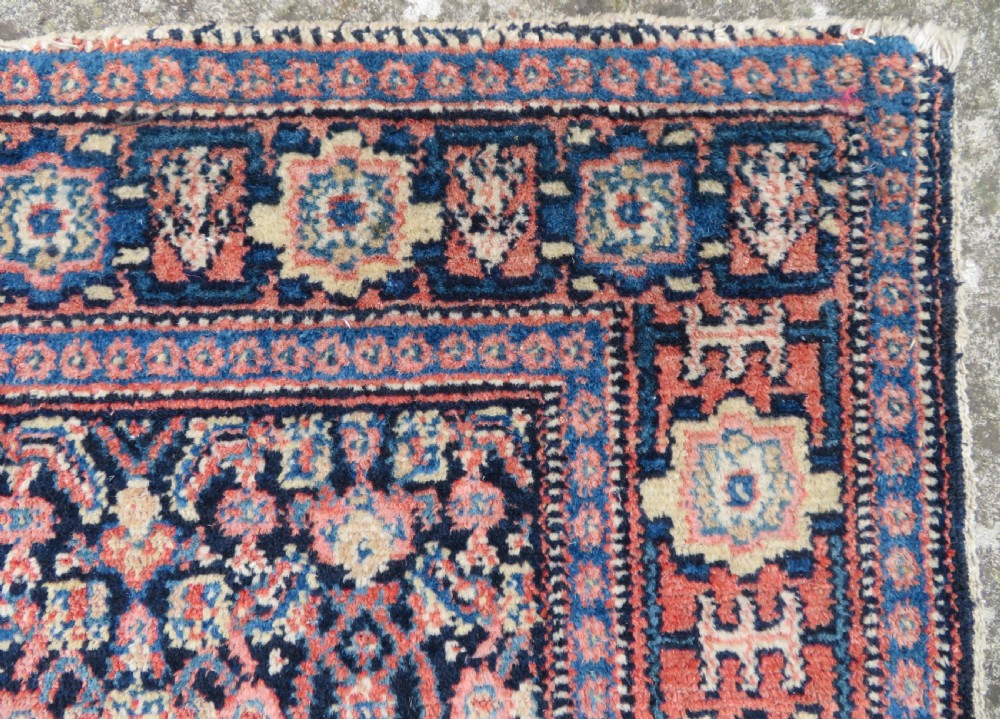 An antique Kurdish Senneh hand-knotted wool rug featuring a central stepped diamond medallion surrounded by detailed repeating motifs in navy blue, terracotta red, and cream. View X