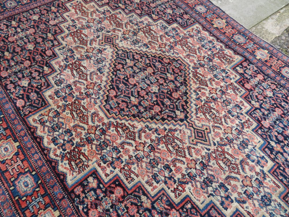 An antique Kurdish Senneh hand-knotted wool rug featuring a central stepped diamond medallion surrounded by detailed repeating motifs in navy blue, terracotta red, and cream. View XIII