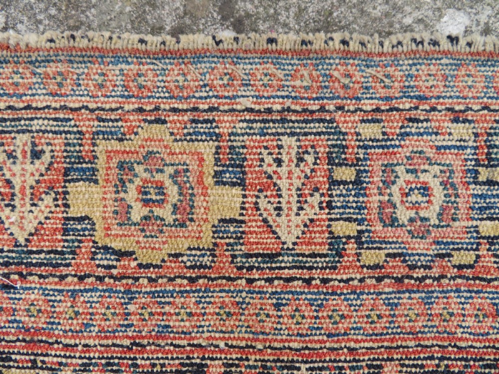 An antique Kurdish Senneh hand-knotted wool rug featuring a central stepped diamond medallion surrounded by detailed repeating motifs in navy blue, terracotta red, and cream. View XVI