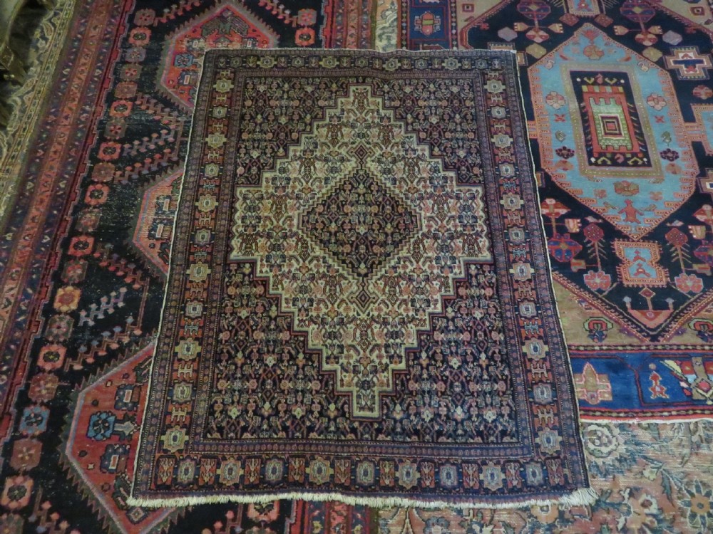 An antique Kurdish Senneh hand-knotted wool rug featuring a central stepped diamond medallion surrounded by detailed repeating motifs in navy blue, terracotta red, and cream. View XVIII