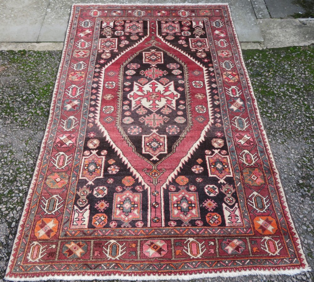 A rectangular hand-knotted North West Persian Kurdish wool rug featuring a central crimson diamond medallion surrounded by geometric motifs on a dark brown field with decorative borders.