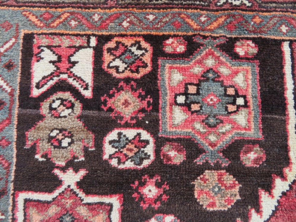 A rectangular hand-knotted North West Persian Kurdish wool rug featuring a central crimson diamond medallion surrounded by geometric motifs on a dark brown field with decorative borders. View V