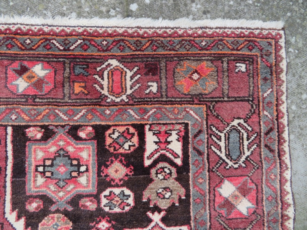 A rectangular hand-knotted North West Persian Kurdish wool rug featuring a central crimson diamond medallion surrounded by geometric motifs on a dark brown field with decorative borders. View X