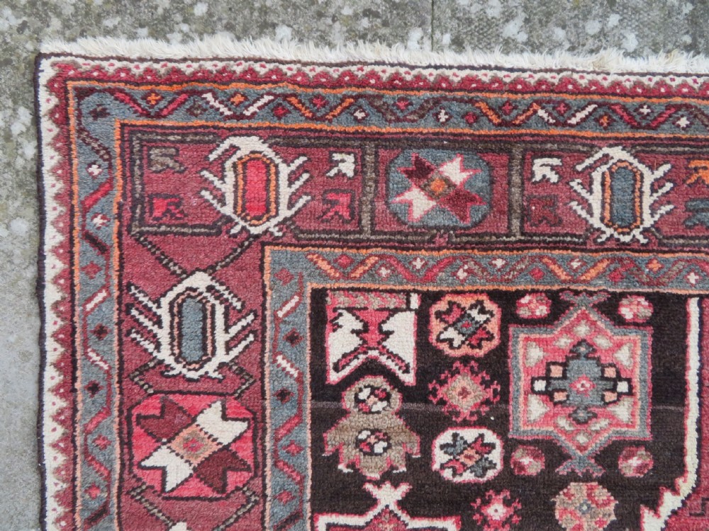A rectangular hand-knotted North West Persian Kurdish wool rug featuring a central crimson diamond medallion surrounded by geometric motifs on a dark brown field with decorative borders. View XI