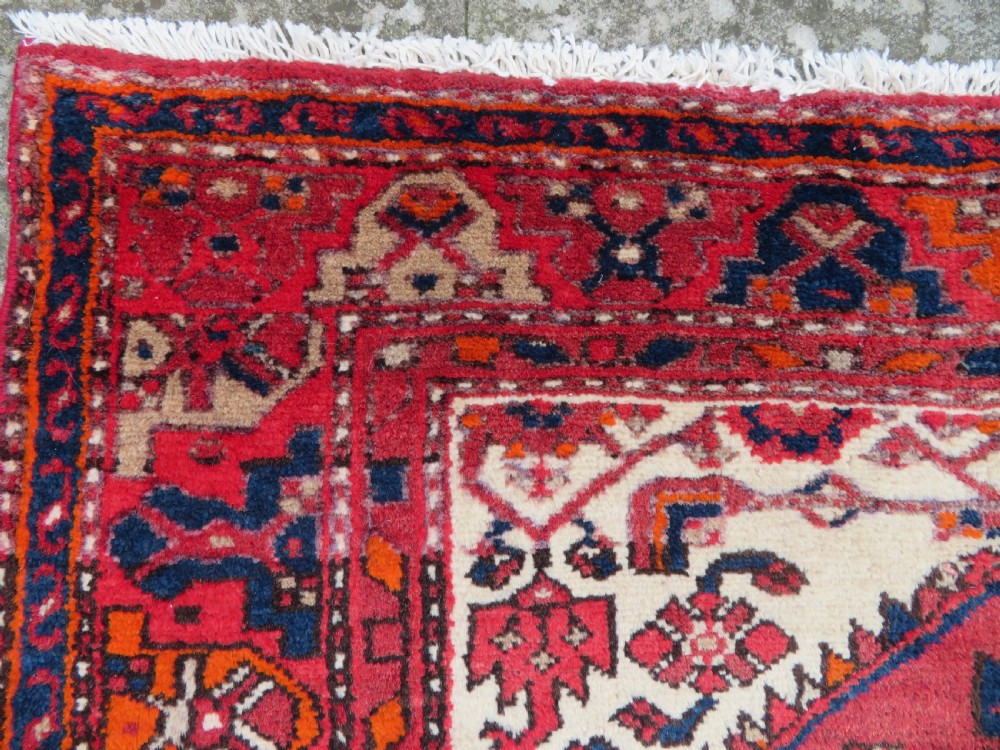 An antique Persian Malayer hand-knotted wool rug featuring a red central field, stepped navy medallion, ivory spandrels, and geometric floral borders. View IX