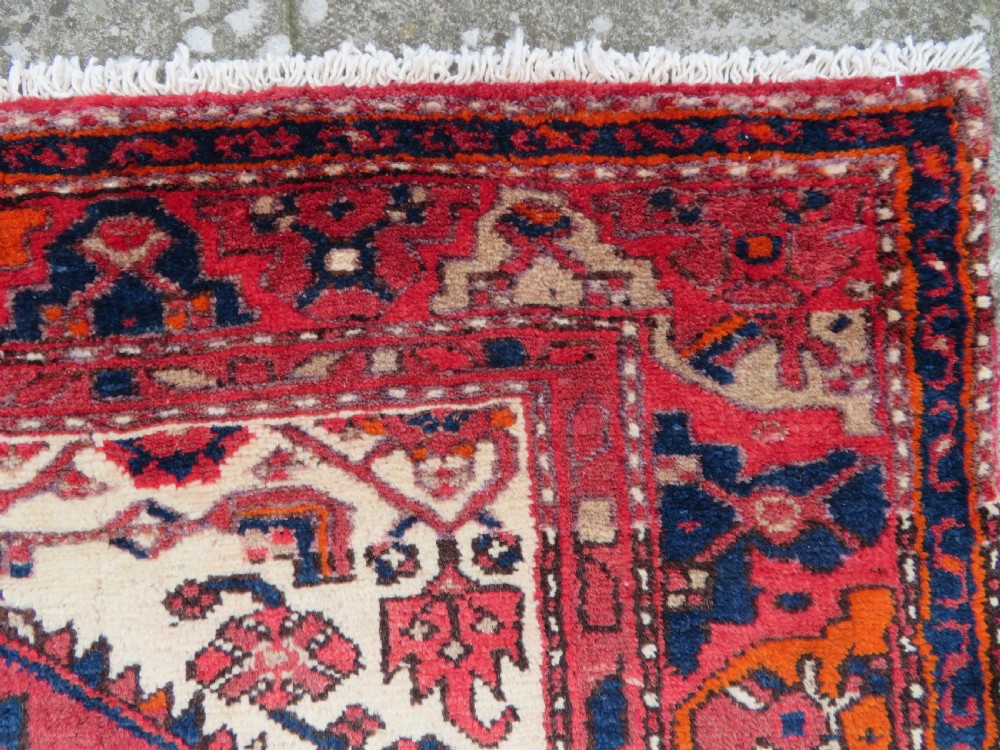 An antique Persian Malayer hand-knotted wool rug featuring a red central field, stepped navy medallion, ivory spandrels, and geometric floral borders. View X