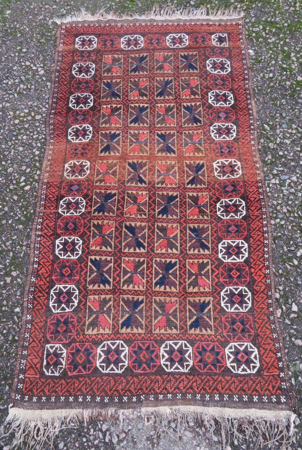 An antique Baluch wool rug featuring a repeating grid pattern of geometric motifs in rust red, dark blue, brown, and ivory accents with a fringed border.