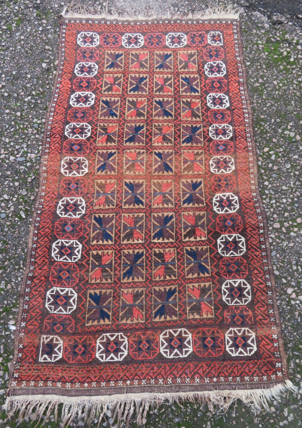 An antique Baluch wool rug featuring a repeating grid pattern of geometric motifs in rust red, dark blue, brown, and ivory accents with a fringed border. View II
