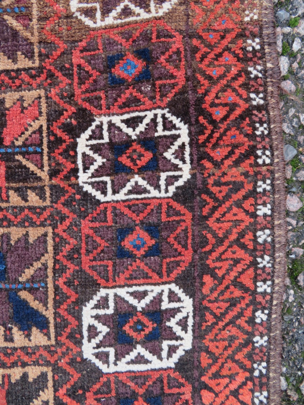 An antique Baluch wool rug featuring a repeating grid pattern of geometric motifs in rust red, dark blue, brown, and ivory accents with a fringed border. View III