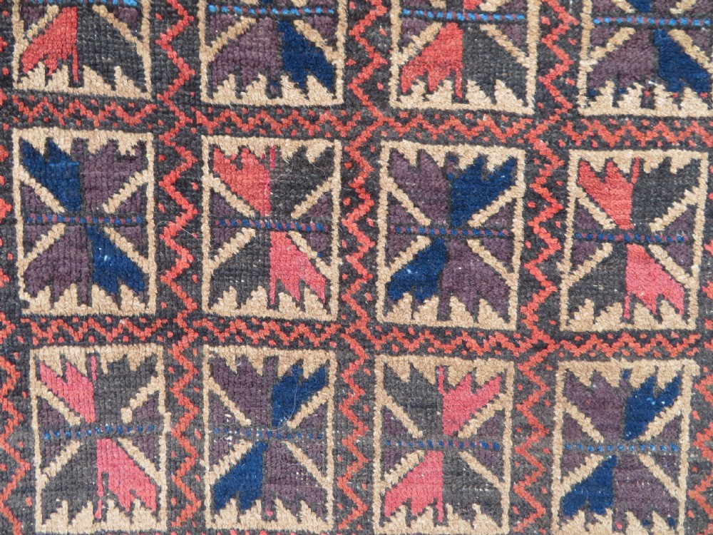 An antique Baluch wool rug featuring a repeating grid pattern of geometric motifs in rust red, dark blue, brown, and ivory accents with a fringed border. View V