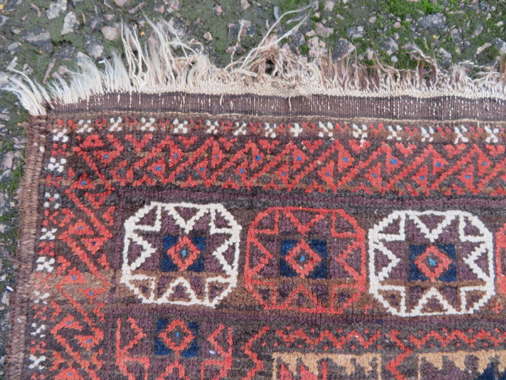 An antique Baluch wool rug featuring a repeating grid pattern of geometric motifs in rust red, dark blue, brown, and ivory accents with a fringed border. View VI
