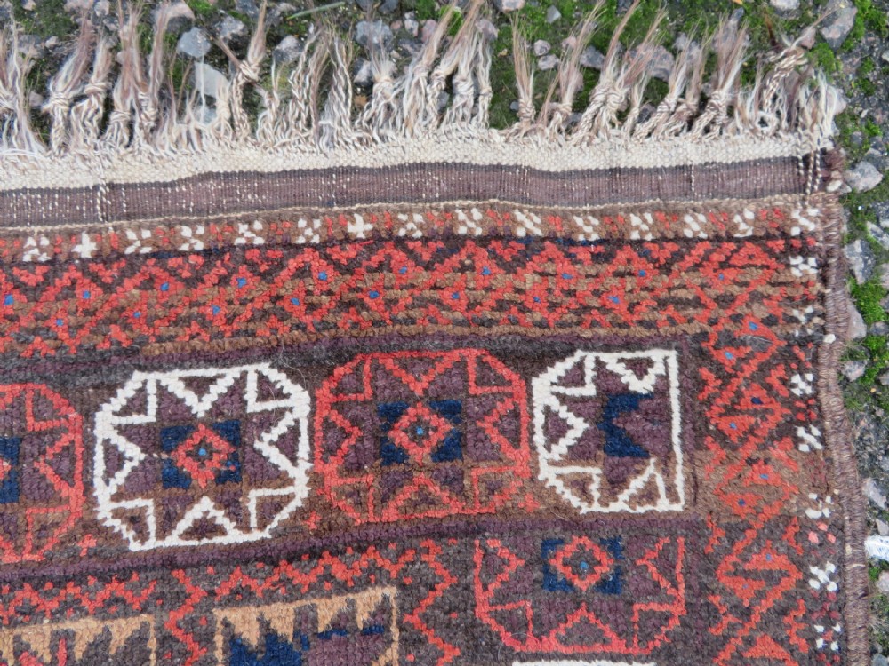 An antique Baluch wool rug featuring a repeating grid pattern of geometric motifs in rust red, dark blue, brown, and ivory accents with a fringed border. View VII