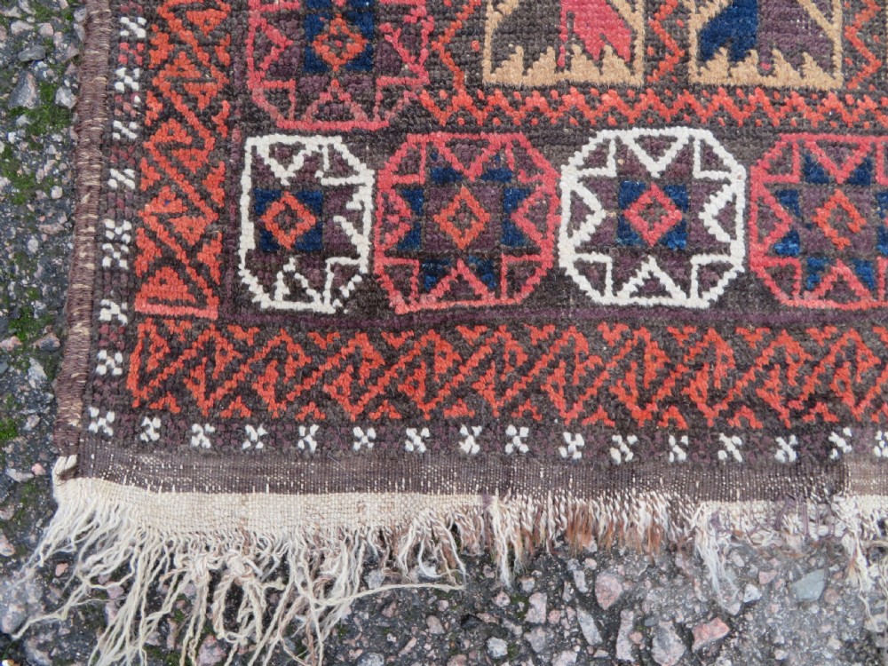 An antique Baluch wool rug featuring a repeating grid pattern of geometric motifs in rust red, dark blue, brown, and ivory accents with a fringed border. View VIII